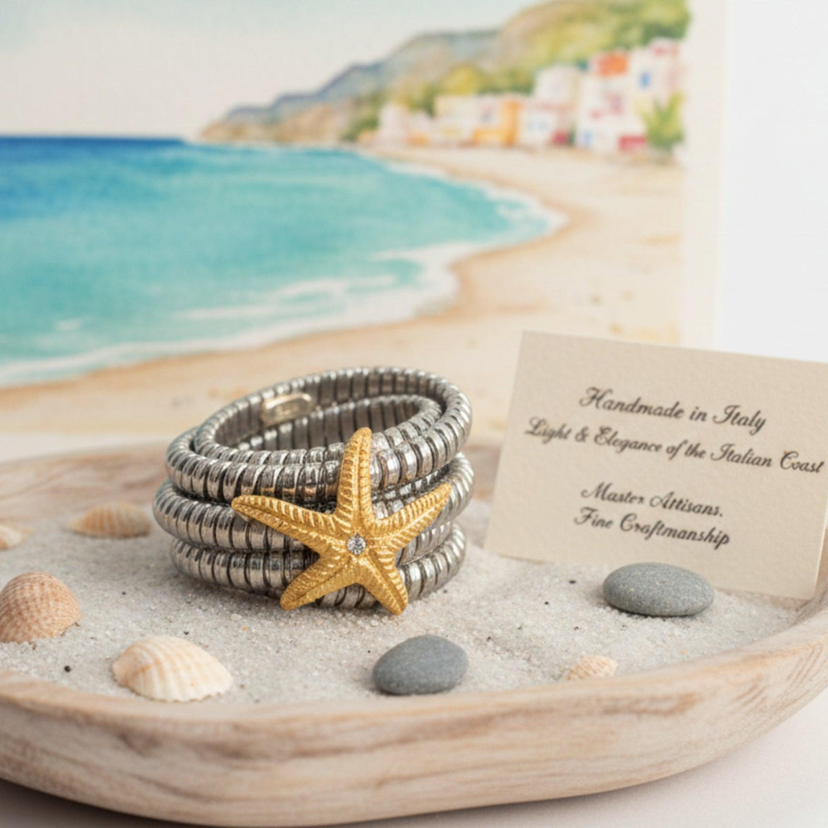 Anello in acciaio con stella marina dorata, su un vassoio con sabbia, conchiglie e sassi, con il mare sullo sfondo