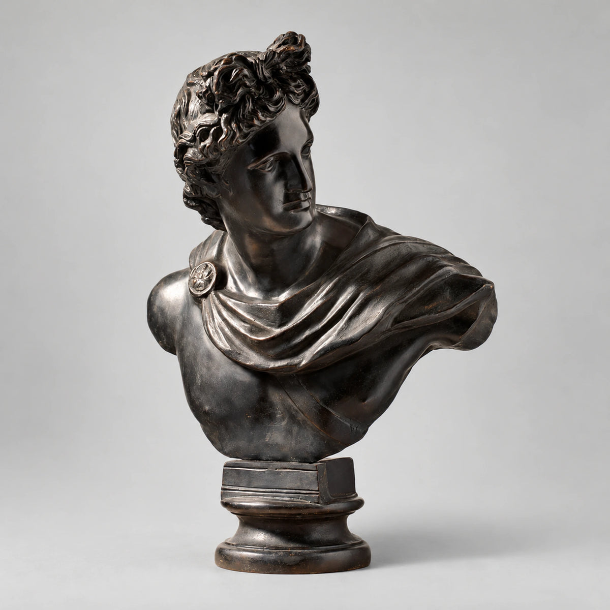 Busto di Apollo del Belvedere in bronzo scuro, alto 70 cm, con drappeggio e base decorativa su fondo grigio chiaro.