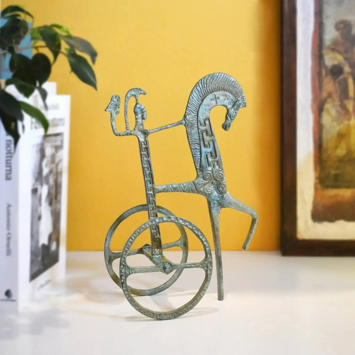 Bronze statuette of a Greek chariot with green patina, stylized horse, and decorative wheels on a white base.