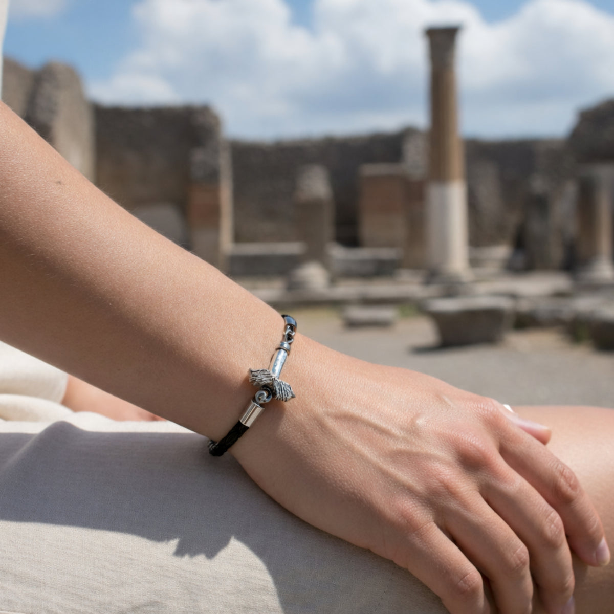 Bracciale in filo nero intrecciato con fallo alato in argento, indossato al polso davanti a rovine con colonne