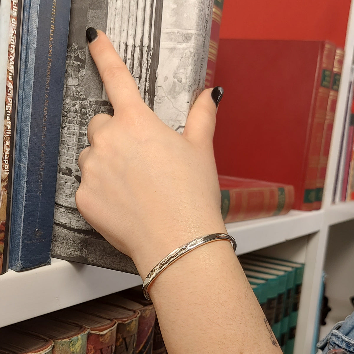 Bracciale in argento con serpente in rilievo, indossato al polso davanti a una libreria