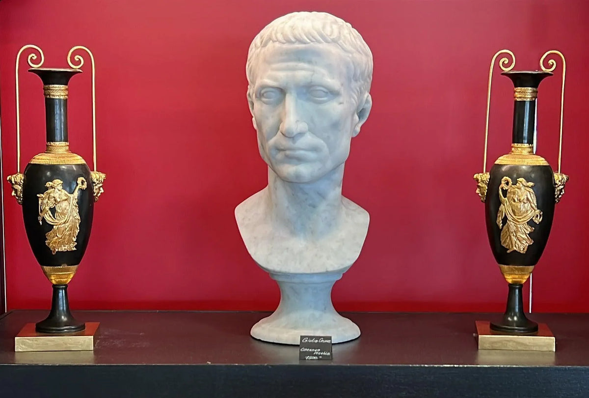 Busto di Giulio Cesare in marmo bianco di Carrara,  vista frontale su un piano di legno  vicino a vasi dorati.