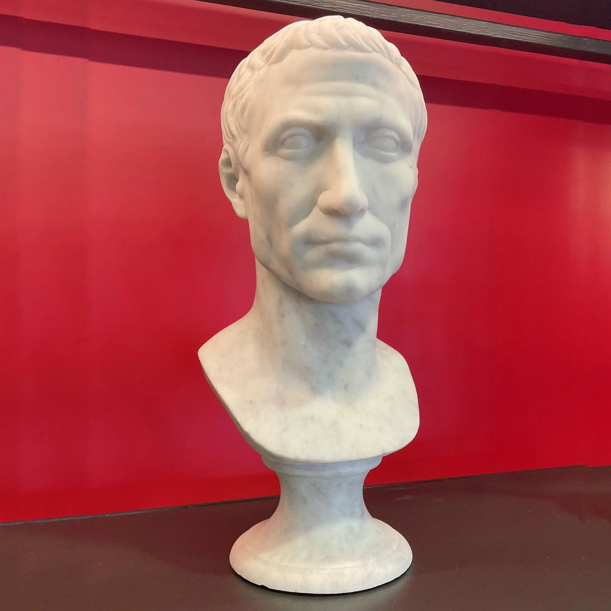 Busto di Giulio Cesare in marmo bianco di Carrara,  vista frontale su fondo rosso.