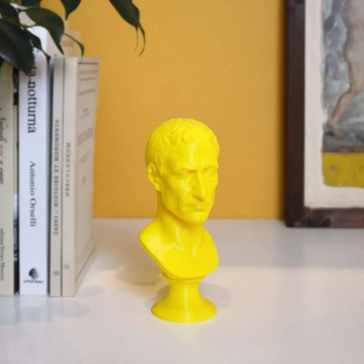 Busto di Giulio Cesare stampato in 3D giallo su una mensola accanto a libri e parete gialla.