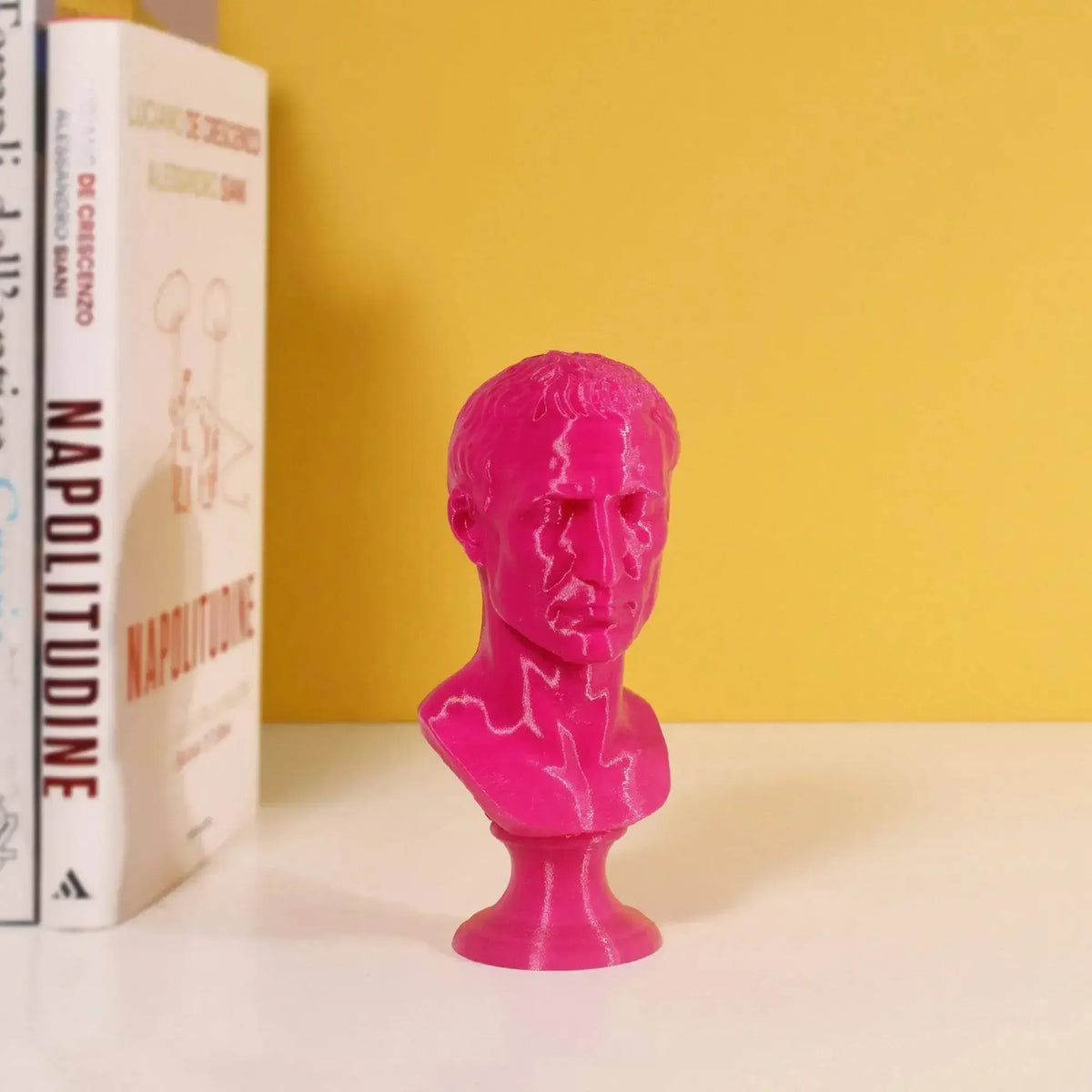 Busto di Giulio Cesare stampato in 3D magenta su una mensola accanto a libri e parete gialla.