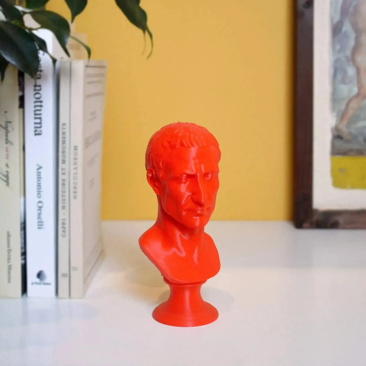 Busto di Giulio Cesare stampato in 3D rosso su una mensola accanto a libri e parete gialla.