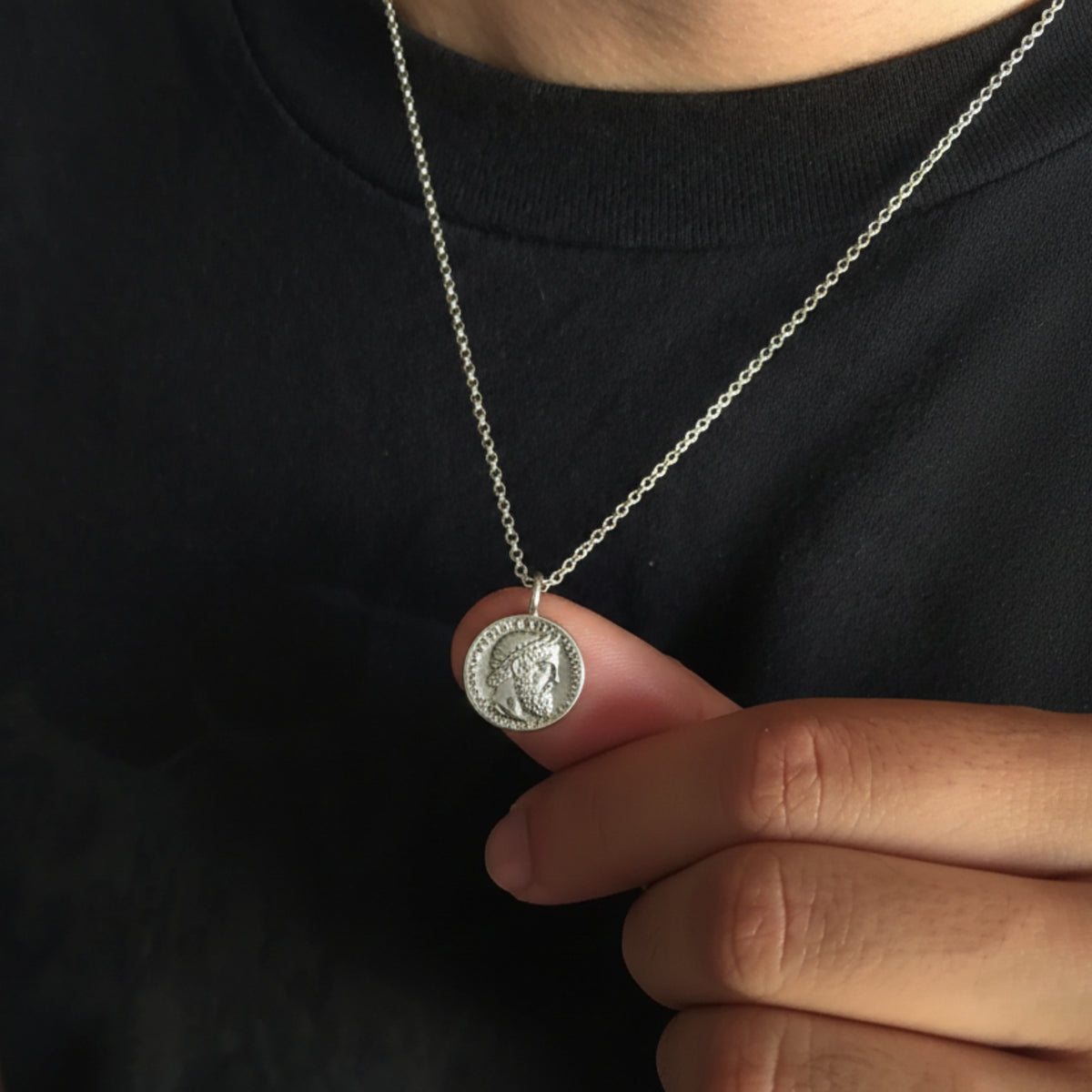 Ciondolo moneta di Naxos in argento, tenuto tra le dita sopra una maglia nera