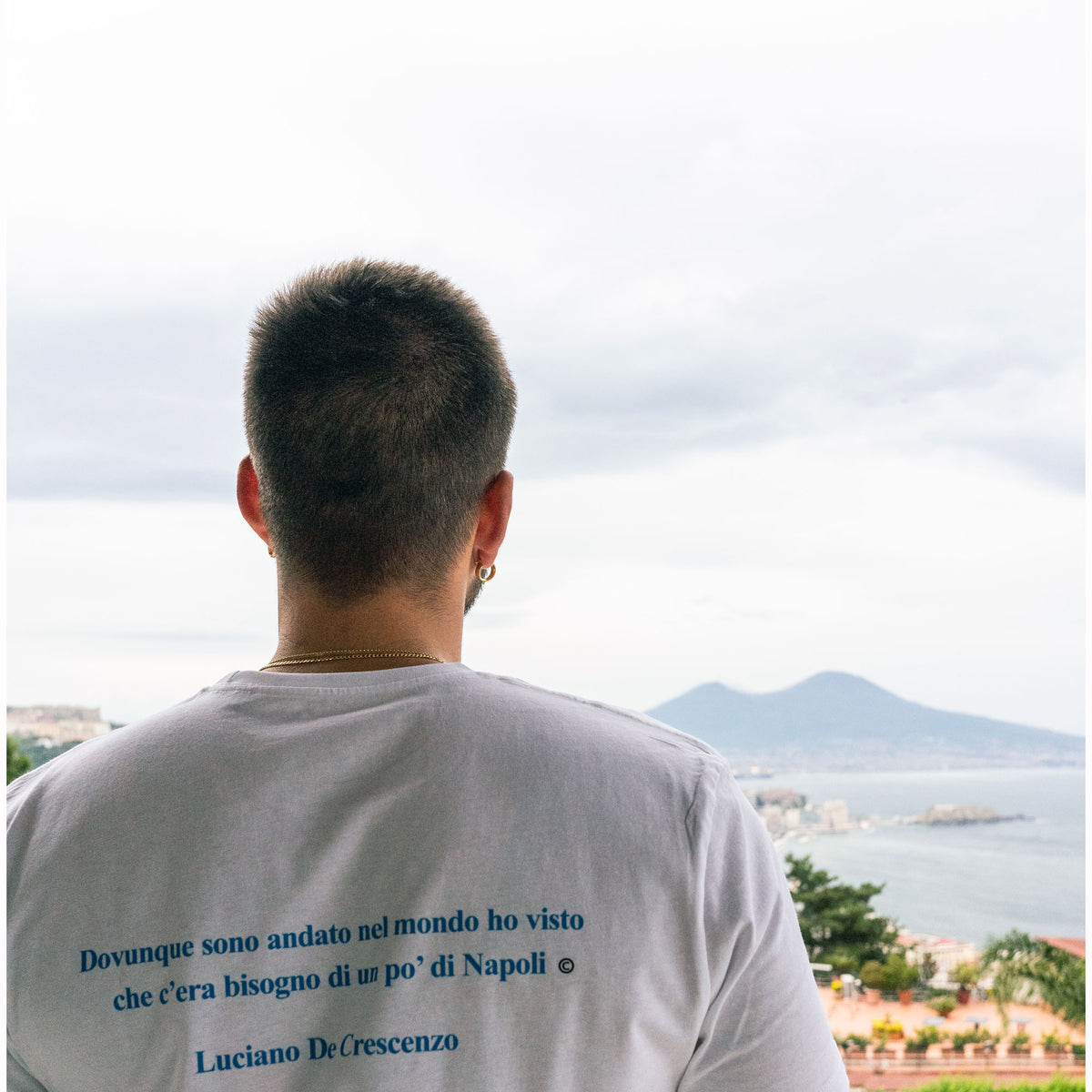 Maglietta bianca indossata di spalle, con citazione di Luciano De Crescenzo su Napoli e panorama sullo sfondo