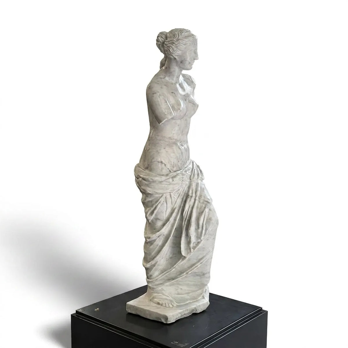 Venere di Milo in marmo di Carrara, vista di profilo su piedistallo nero e fondo bianco.