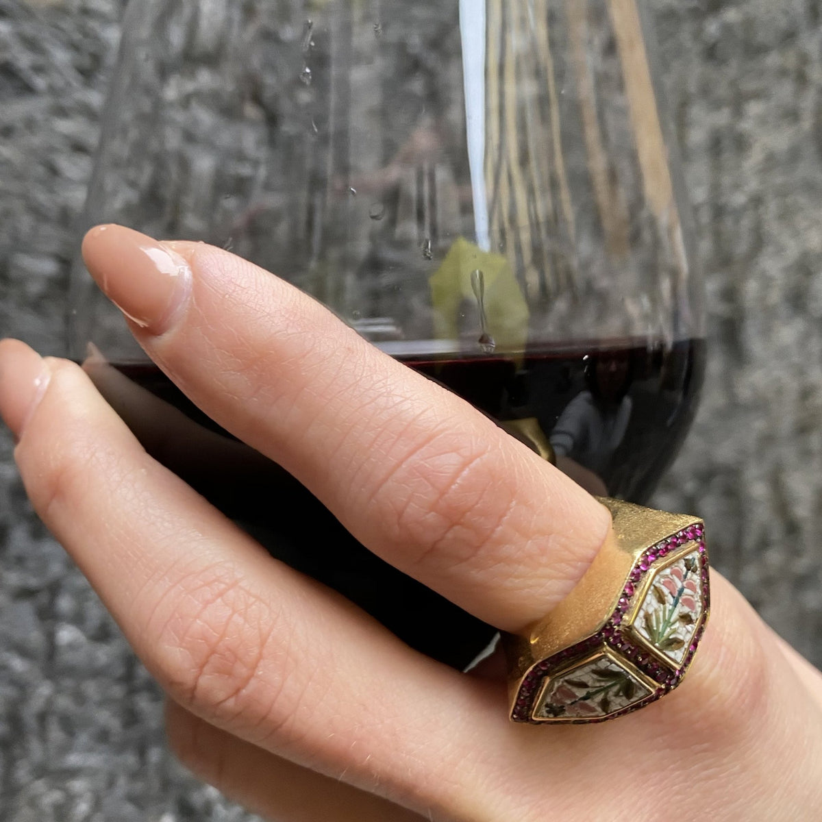Dettaglio dell'anello Hibiscus in oro con micromosaico floreale e rubini, indossato mentre la mano tiene un calice