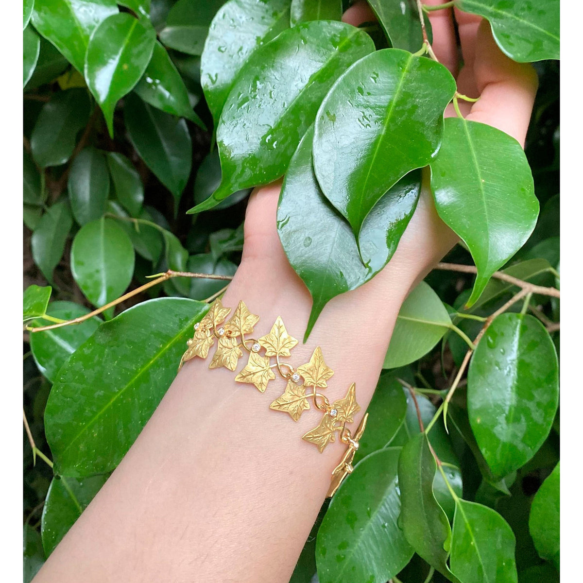 Bracciale in oro con foglie di edera e brillanti, indossato al polso tra foglie verdi