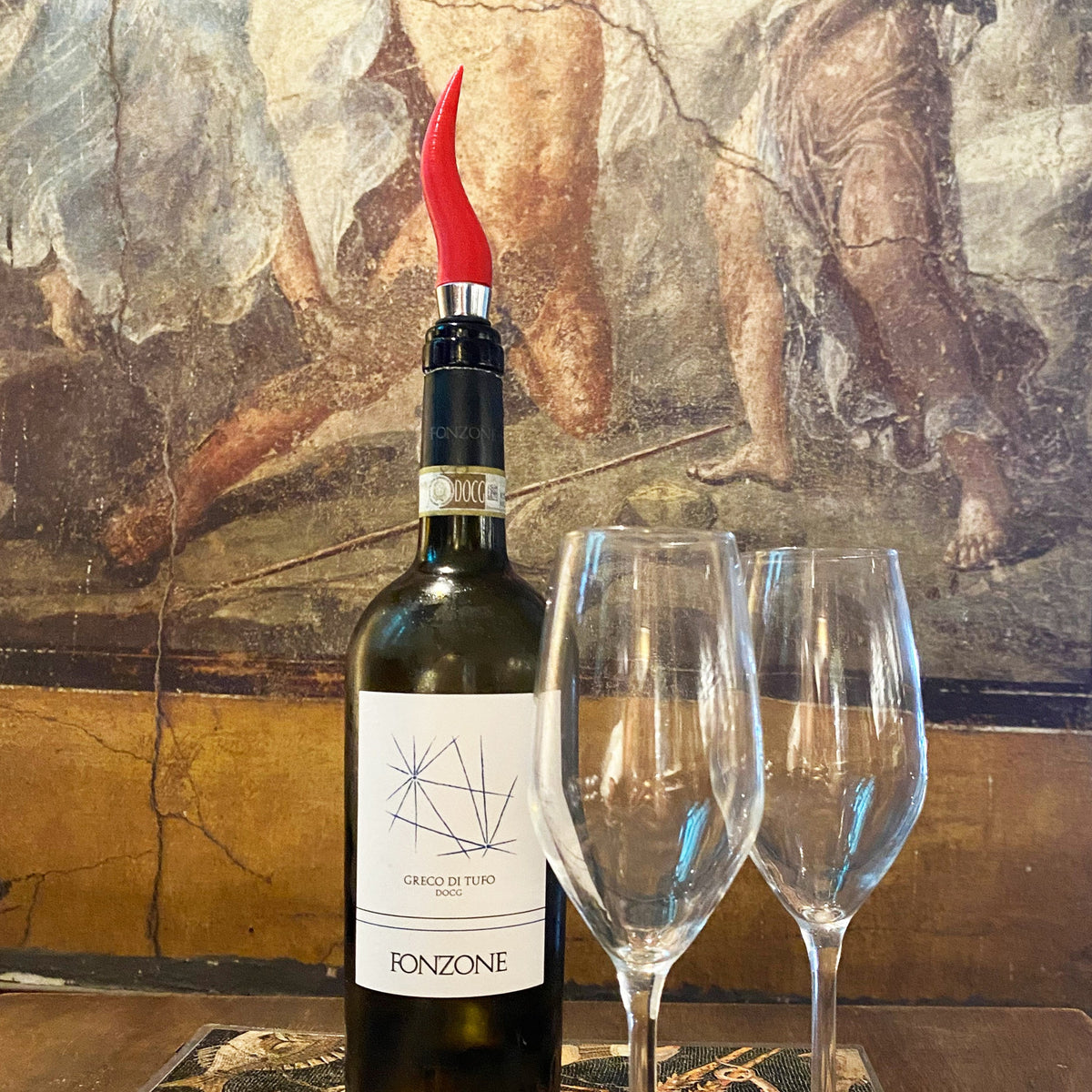 Tappo a corno napoletano rosso su una bottiglia accanto a due calici, davanti a una parete dipinta.